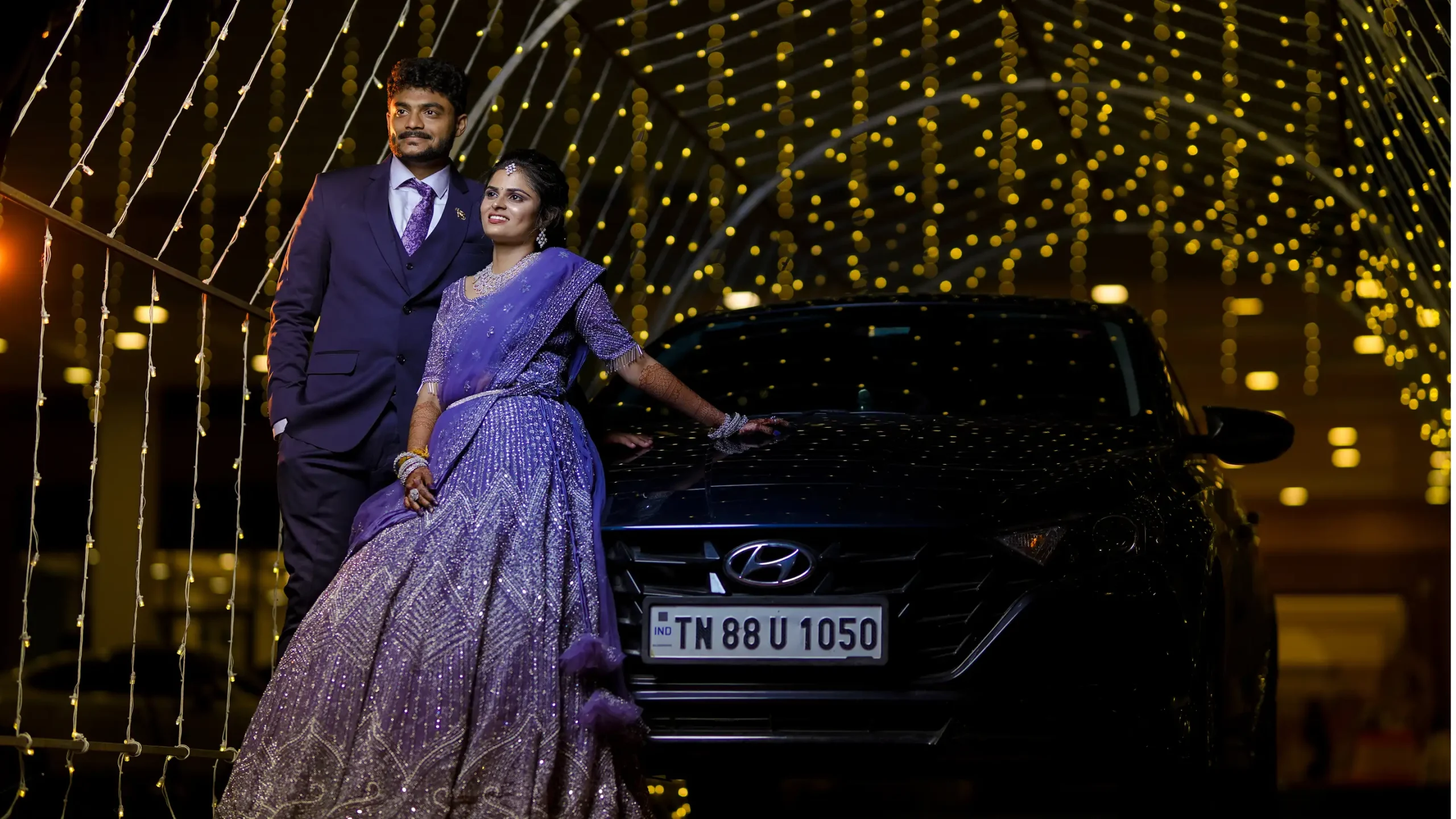Yuvi Studio captures a bride in a royal purple lehenga and a groom in a deep blue suit standing beside a car, illuminated by golden background lights. 🏢 Yuvi Studio Fotography & Film Making 📷 Best Outdoor Wedding PhotoShoot 📍 Velayuthampalayam, Karur