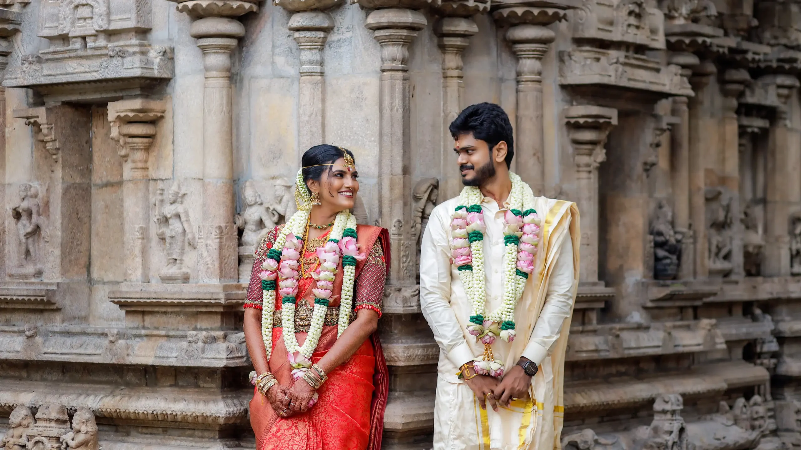Indian bride and groom in white and red traditional attire standing in front of multiple ornate stone temple pillars.🏢 Yuvi Studio Fotography & Film Making 📷 Best Wedding Photographers 📍 Velayuthampalayam, Karur