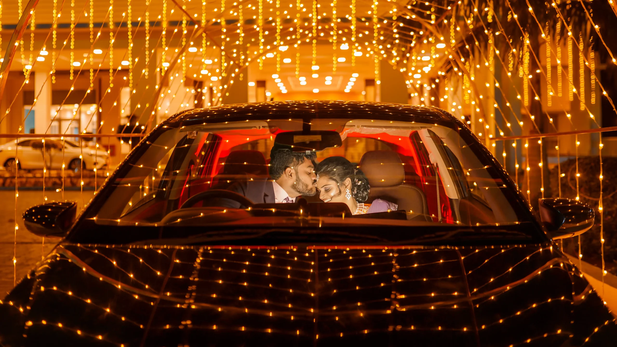 A newlywed groom kisses his smiling bride inside a car, surrounded by glowing lights in the background 🏢 Yuvi Studio Fotography & Film Making 📷 Best Wedding Photographers 📍 Velayuthampalayam, Karur