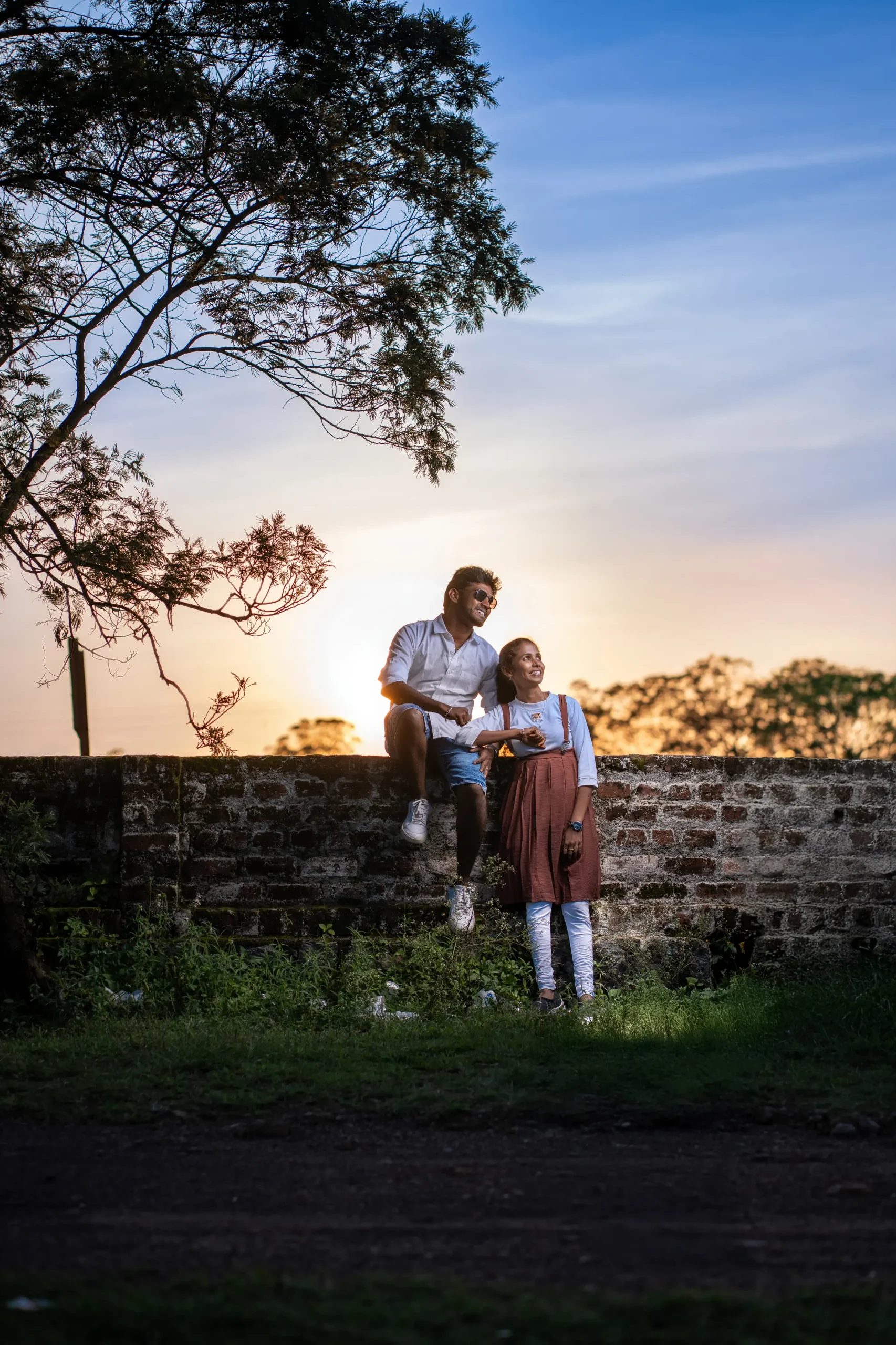 Bride standing in the lane with groom sitting on wall under sunset near green trees Yuvi Studio candid wedding photography π’ Yuvi Studio Fotography & Film Making π· Best Pre-WeddingPhotoshoot π Velayuthampalayam, Karur