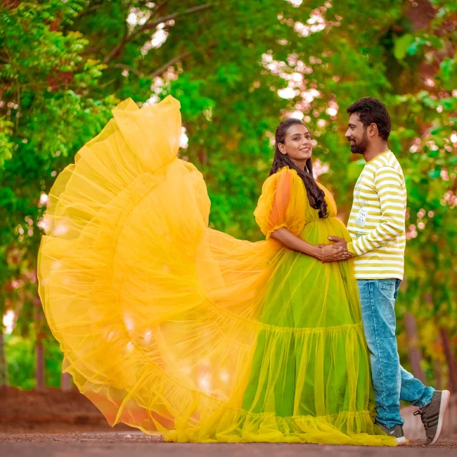 Yuvi Studio captures an expecting couple standing on a road lined with green trees. The mom-to-be, in a yellow and green gown, and dad-to-be, in a yellowish-green striped t-shirt, both hold her baby bump, smiling as he looks at her.π’ Yuvi Studio Fotography & Film Making π· Best outdoor Maternity Photographers π Velayuthampalayam, Karur