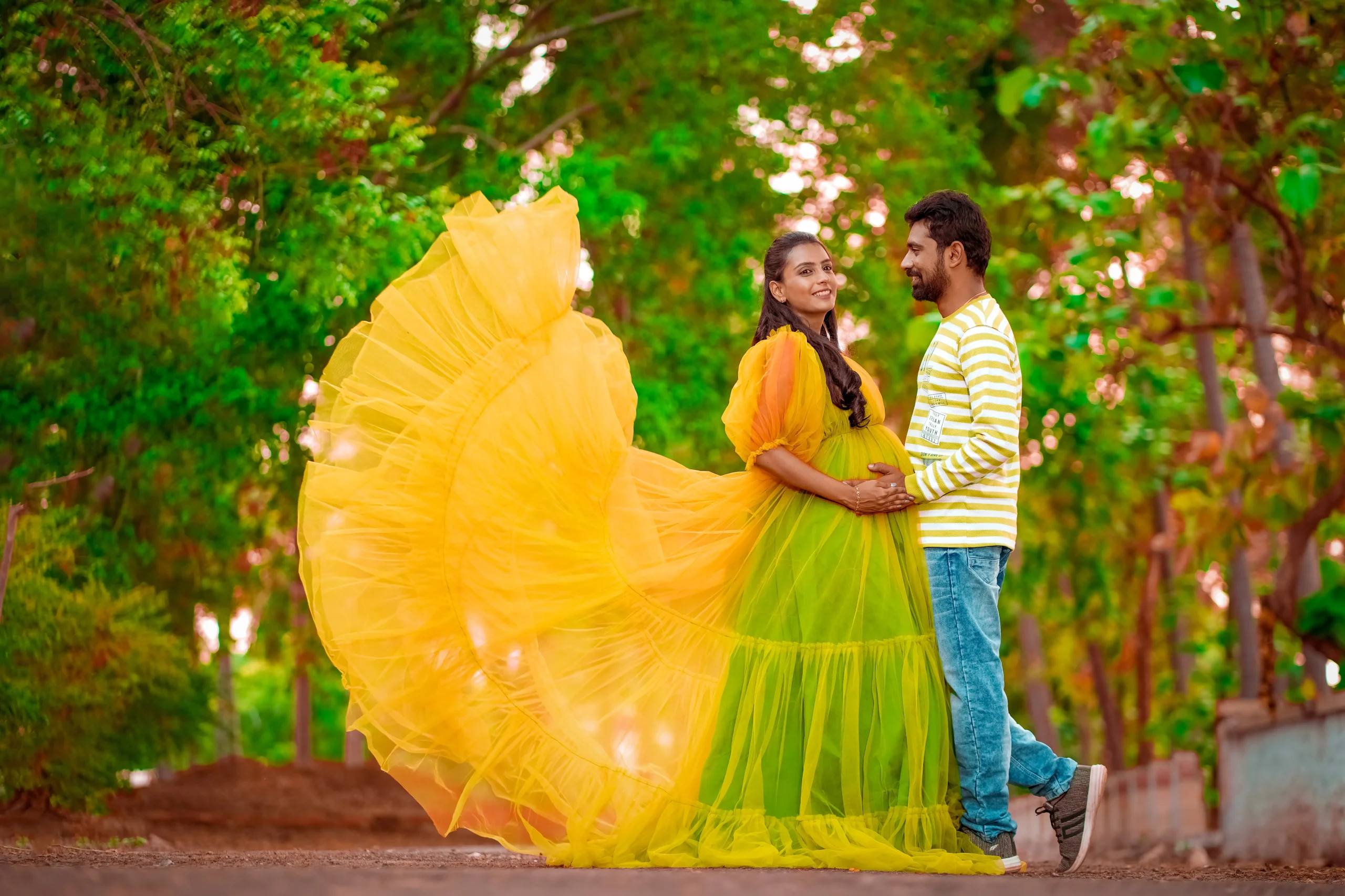 Yuvi Studio captures an expecting couple standing on a road lined with green trees. The mom-to-be, in a yellow and green gown, and dad-to-be, in a yellowish-green striped t-shirt, both hold her baby bump, smiling as he looks at her.π’ Yuvi Studio Fotography & Film Making π· Best outdoor Maternity Photographers π Velayuthampalayam, Karur