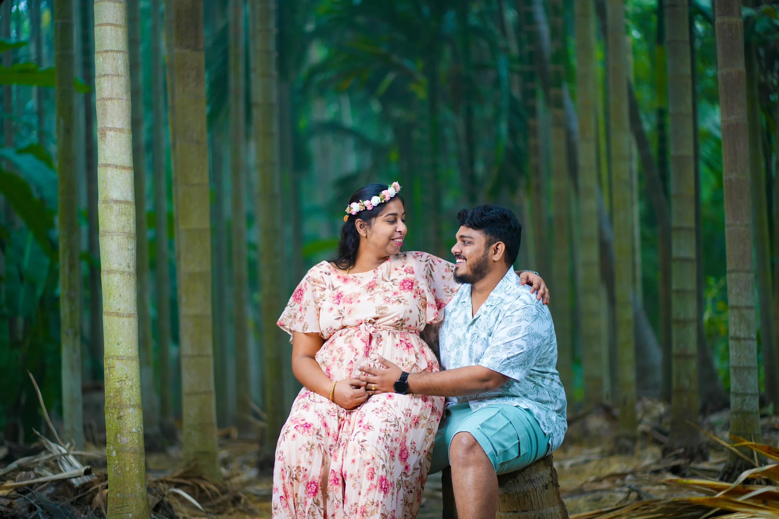 Yuvi Studio captures an expecting couple in a natural setting: mom-to-be in a pink floral dress and crown, sitting on her husband's lap, both smiling at each other as he holds her baby bump. 🏢 Yuvi Studio Fotography & Film Making 📷 Best Natural Maternity Photographers 📍 Velayuthampalayam, Karur