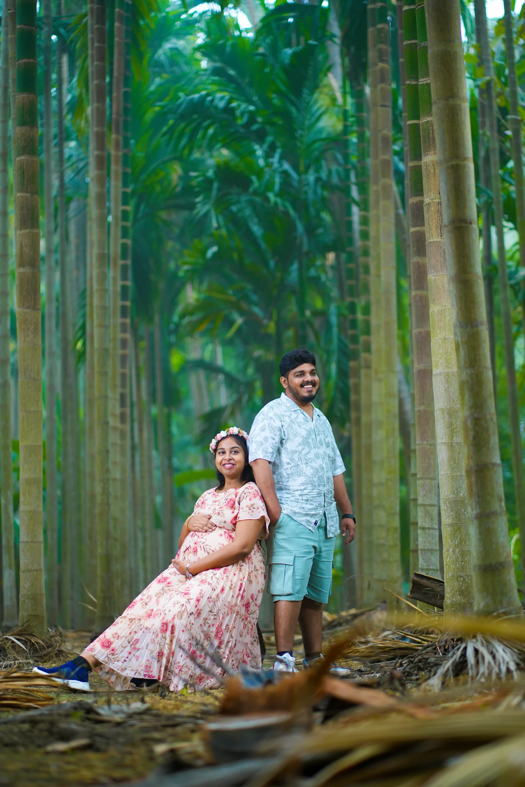 Yuvi Studio maternity photoshoot A mom-to-be in a pink floral dress and crown sits, while her husband stands behind her in a blue shirt 🏢 Yuvi Studio Fotography & Film Making📷 Best Maternity Photographers 📍 Velayuthampalayam, Karur
